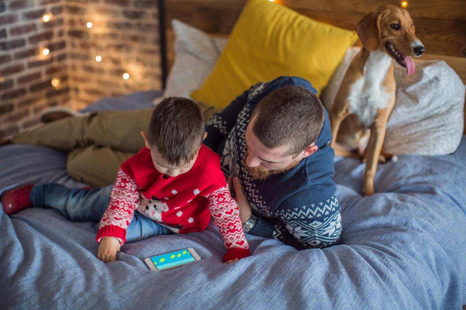 Father and son wearing matching winter sweaters lay on a bed while reading a book.