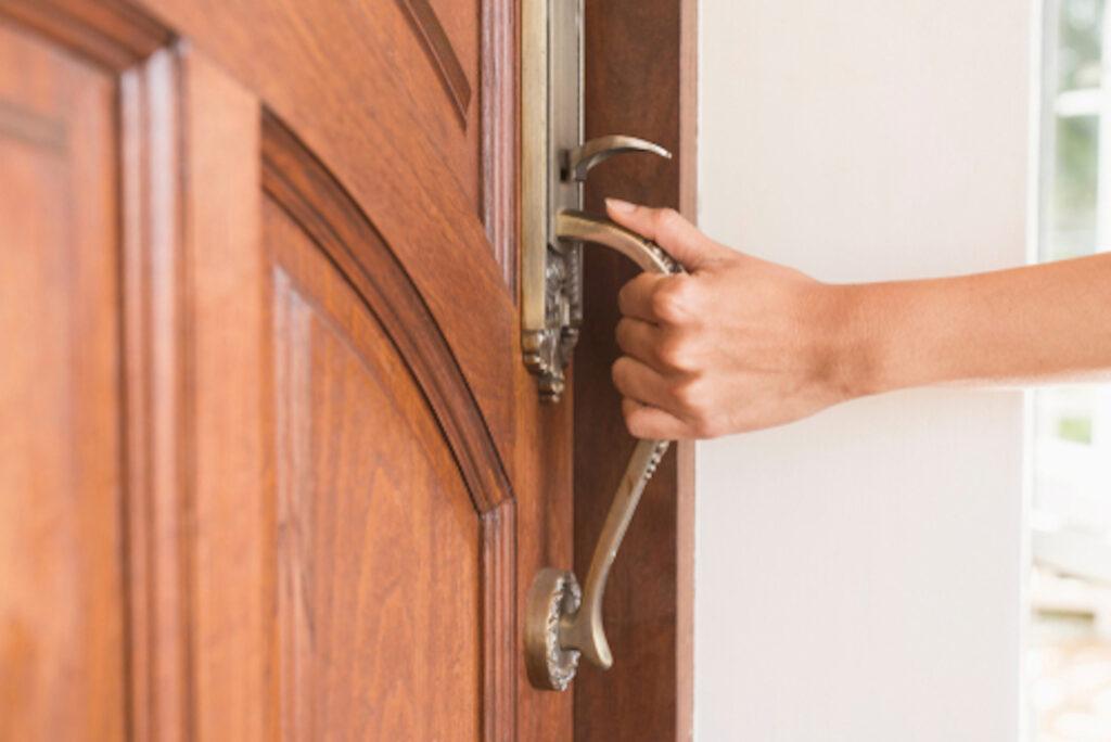 A hand pulls open a wooden front door with a large brass handle.