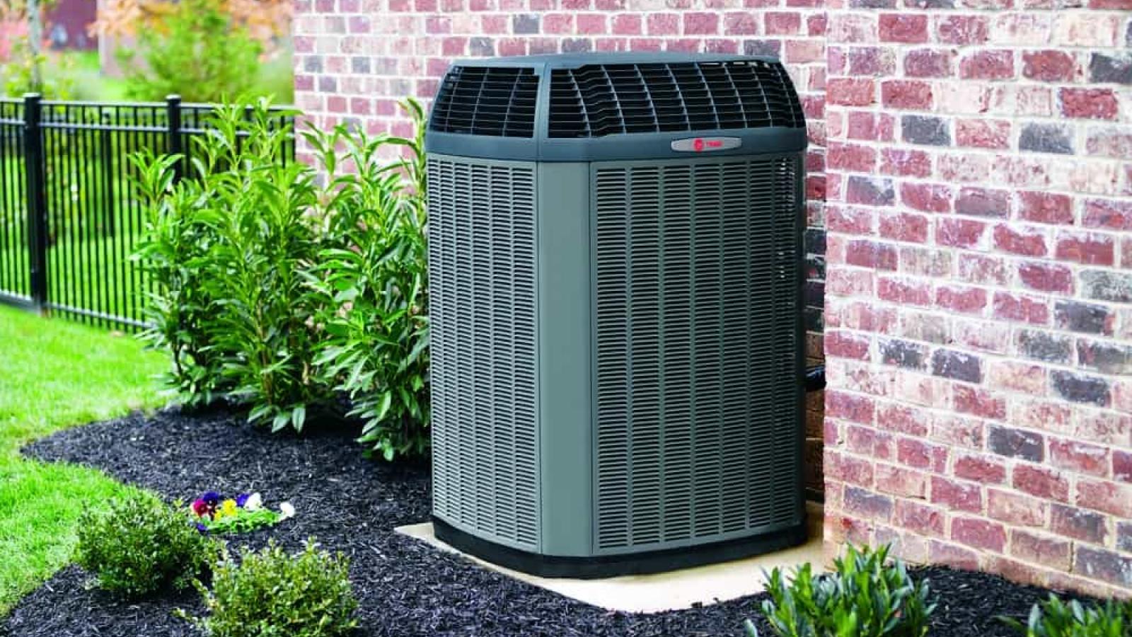 An outdoor Trane HVAC system sits between a red brick wall and green plants.