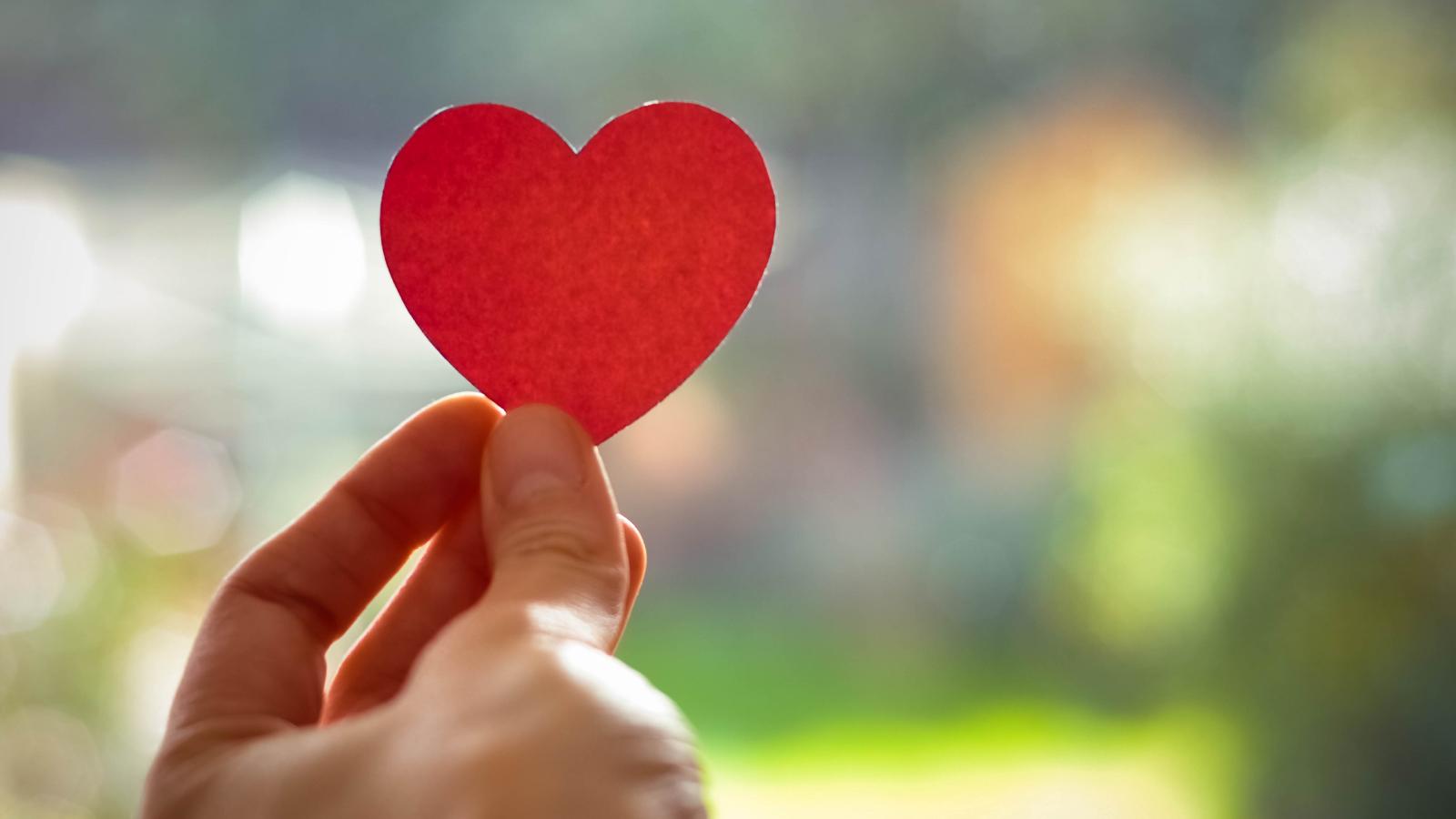 A hand holds up a red paper heart against a blurry background.