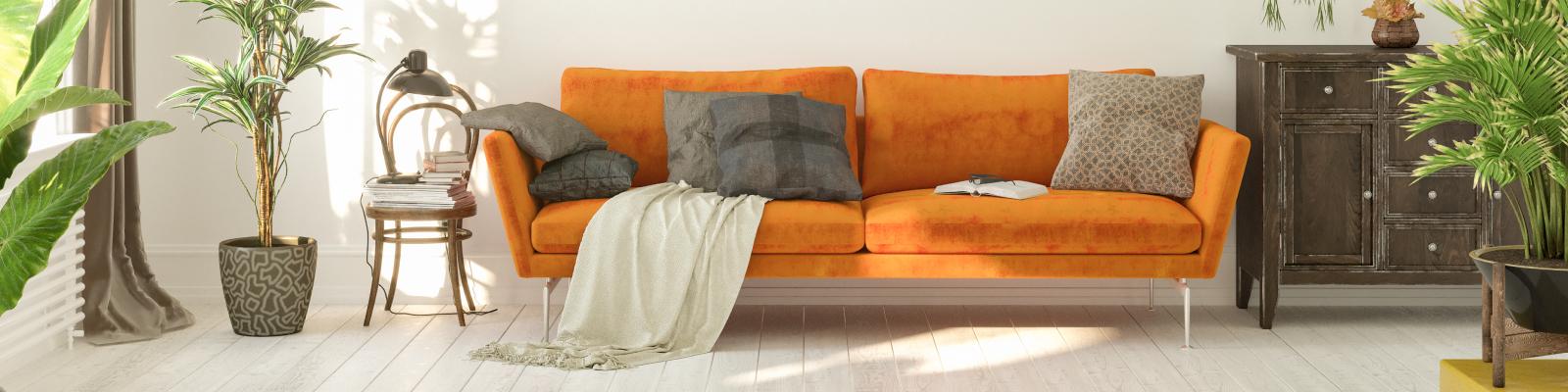 A vibrant orange couch with throw pillows and blankets sits in a living room surrounded by plants.