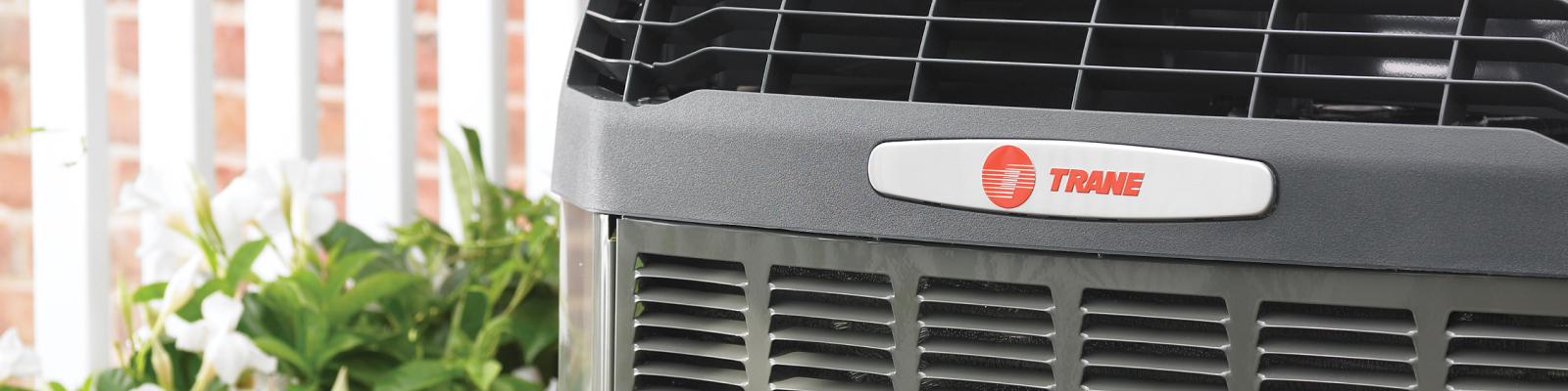 Trane oudoor HVAC system is pictured close up with a white fence and flowers in the background.