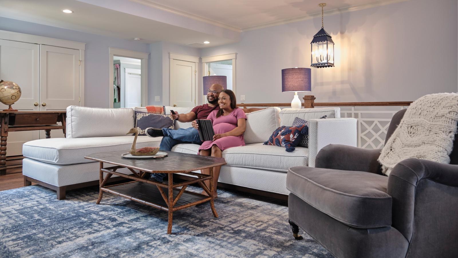 A woman smiles and holds an iPad while sitting next to her husband on the couch watching TV.