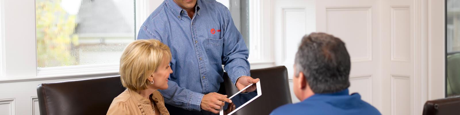 A Trane HVAC dealer sits with a couple at their dining room table and shows them different system options on an iPad.
