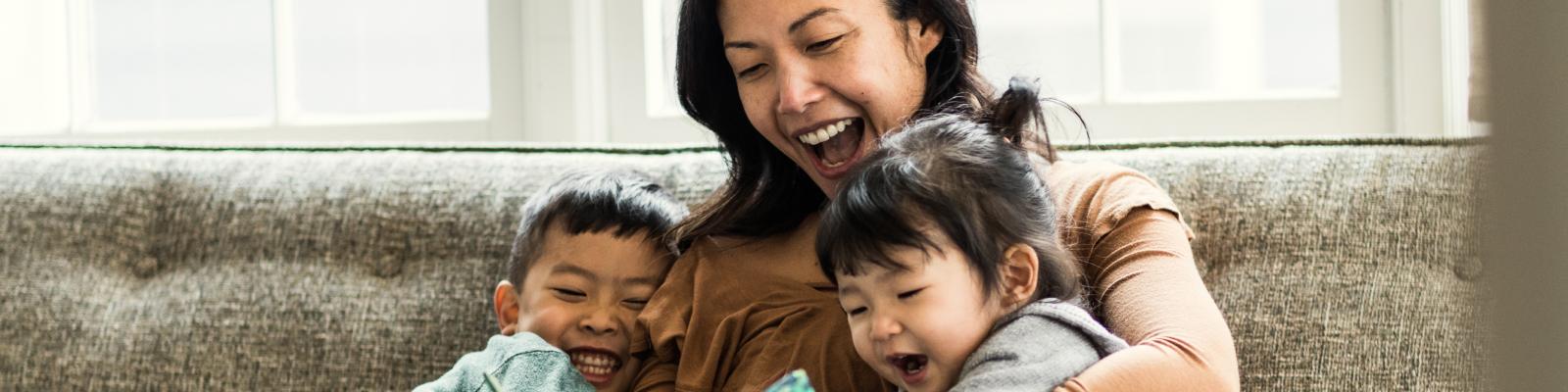 A woman smiles and laughs sitting on a couch with two toddlers in her arms.