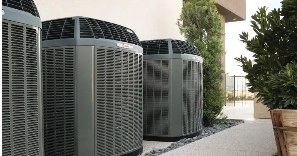 Three outdoor Trane HVAC systems are lined up surrounded by trees and plants.