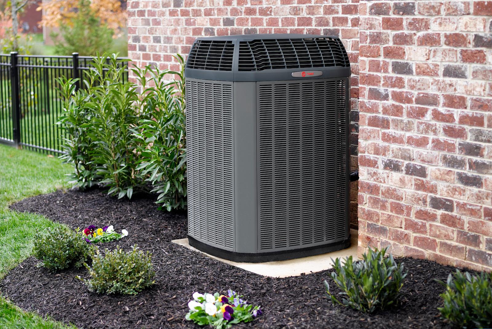 An outdoor Trane HVAC system is nestled between a brick wall and plants.