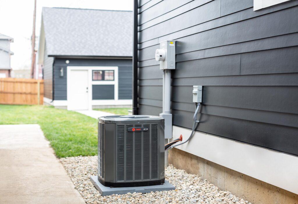 An outdoor Trane HVAC system is plugged into home painted black, with a home painted blue in the background.
