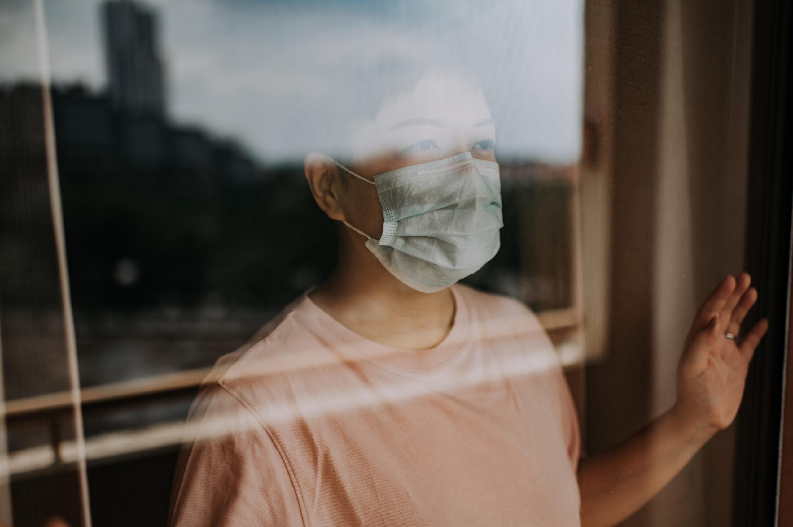 A woman wearing a blue surgical mask and a pink t-shirt looks out a window.