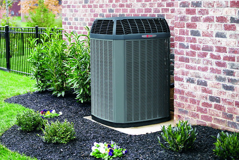 A Trane HVAC system sits on a concrete pad within a well manicured lawn and against a brick wall.
