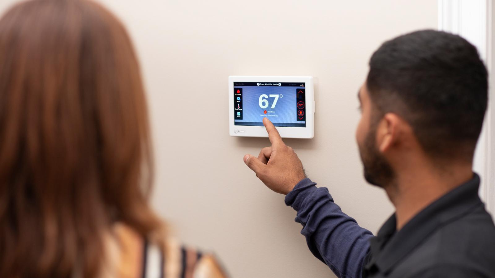 A man shows a woman a Trane smart thermostat reading 67 degrees by pointing at the number on the screen.