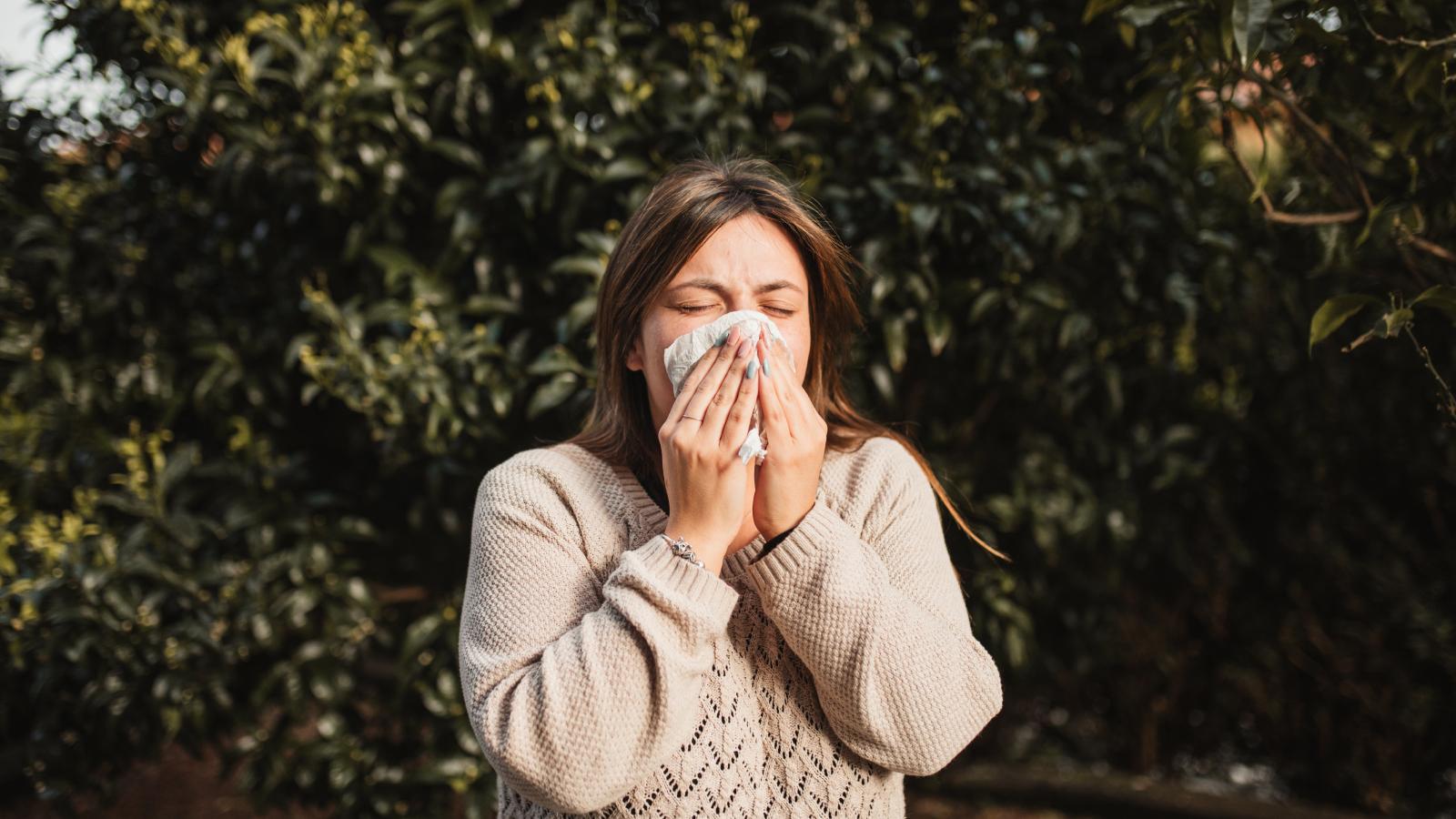 Woman in a tan sweater stands outside sneezing into a tissue.