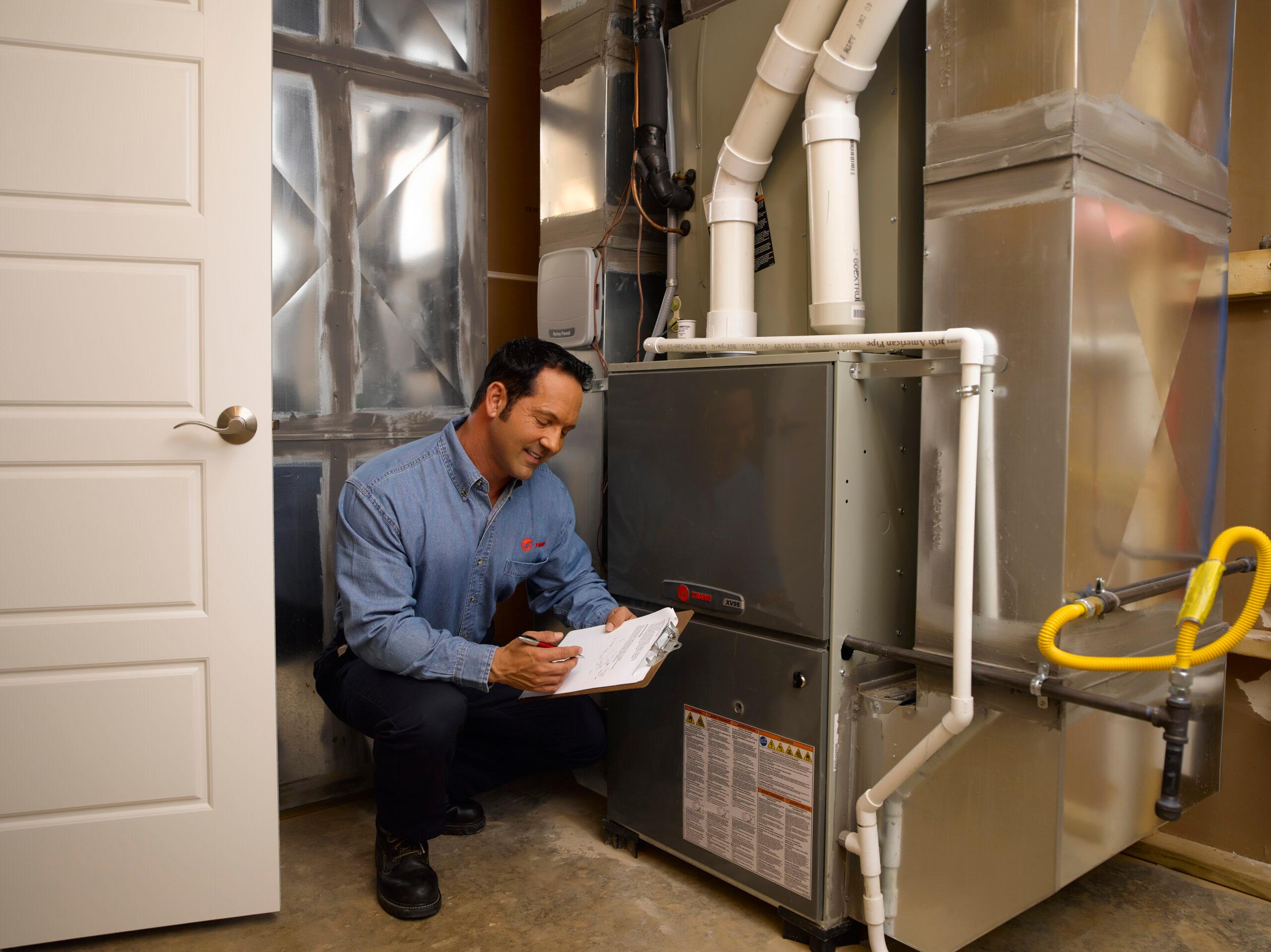Trane HVAC technician holds a clipboard and smiles as he inspects an indoor heating and cooling system.