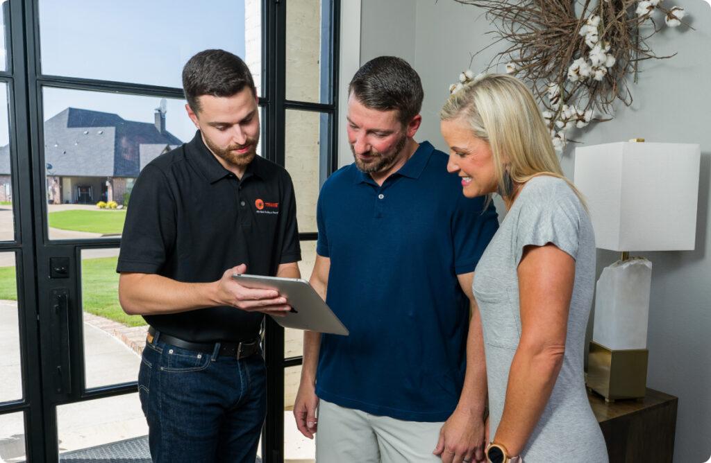 A Trane HVAC technician stands with a man and woman inside a house viewing tablet.