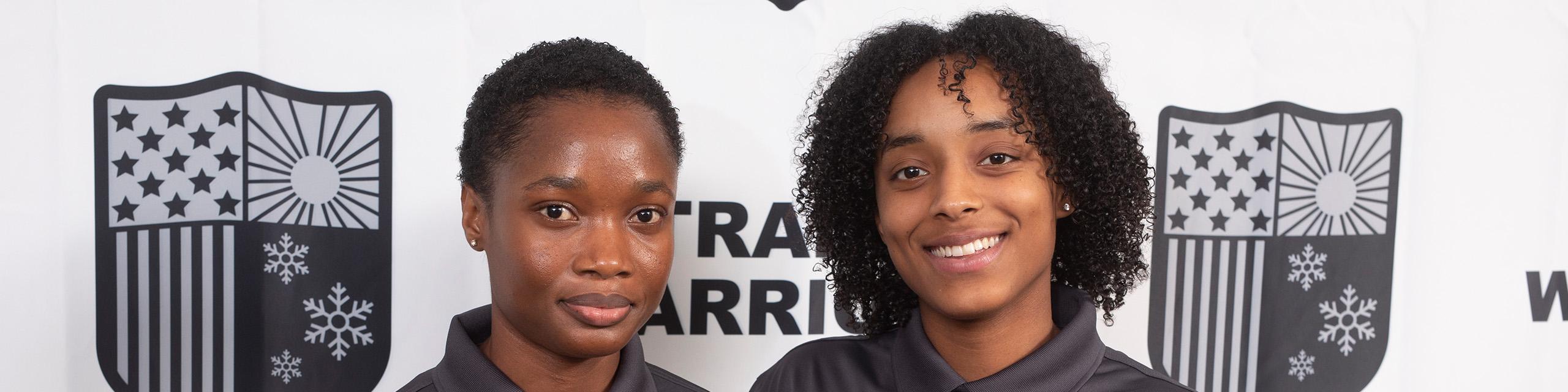 Two young African American women in black shirts are standing and smiling.