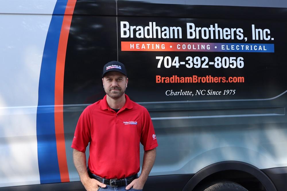 A Bradham Brothers male HVAC tech in a red shirt is standing in front of his service truck.