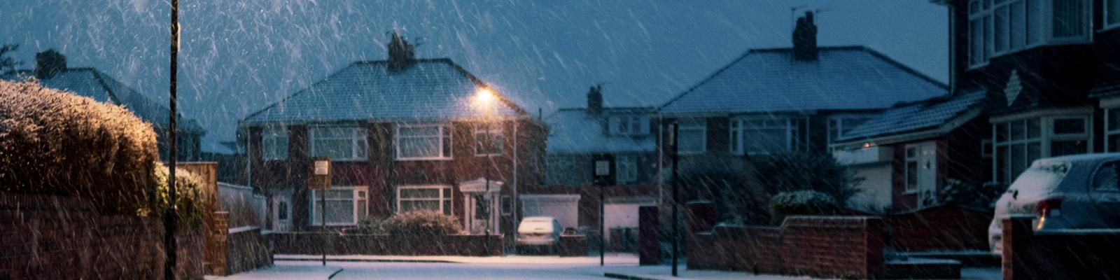 There is a suburban street with some two-story brick houses at dusk in a snow flurry.