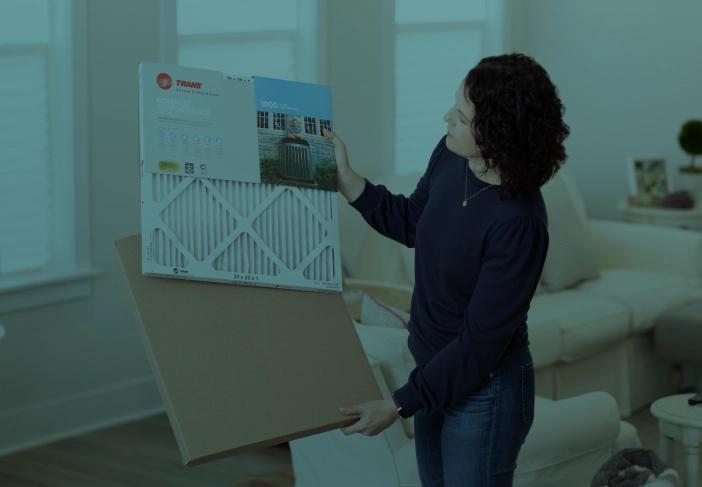 A Caucasian woman in a navy shirt is standing in a a living room holding an air filter.