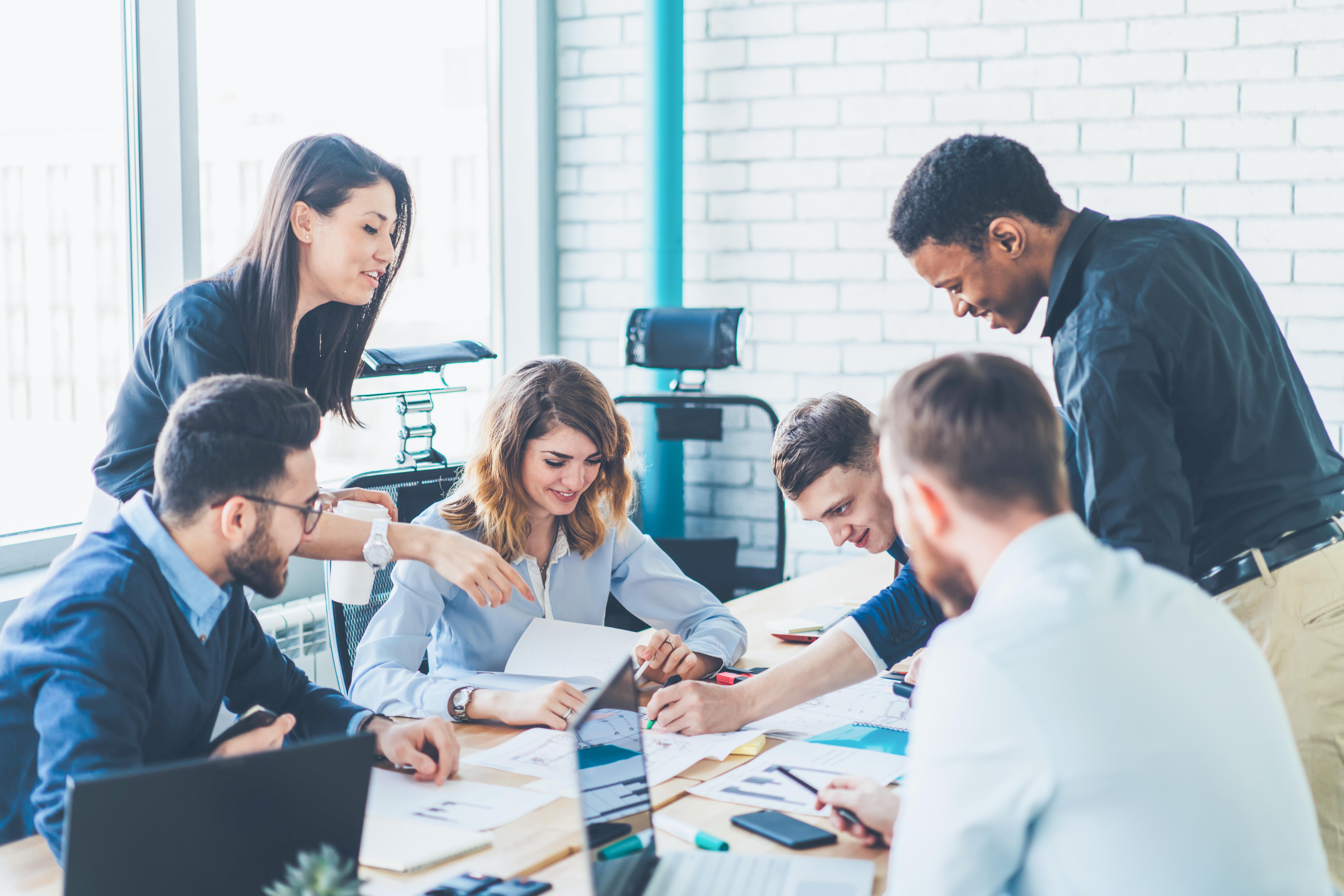 Successful group of male and female business analytics analyzing strategy for startup project during collaborating indoors, positive smiling colleagues enjoying cooperating near office desktop