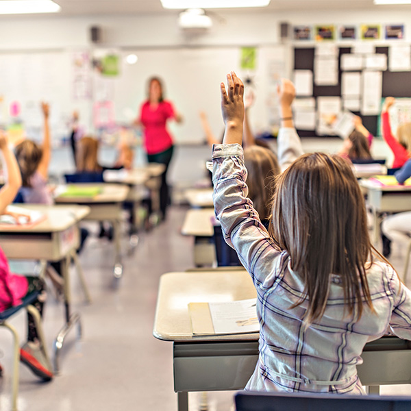 Students raising their hands in a classroom with a teacher at the front of the room